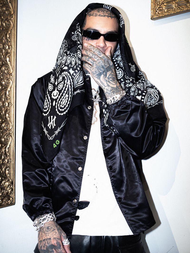 Adult male with tattoos poses indoors wearing a black jacket, bandana hood, t-shirt, bracelets, and sunglasses against a neutral wall.