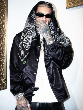 Adult male with tattoos poses indoors wearing a black jacket, bandana hood, t-shirt, bracelets, and sunglasses against a neutral wall.