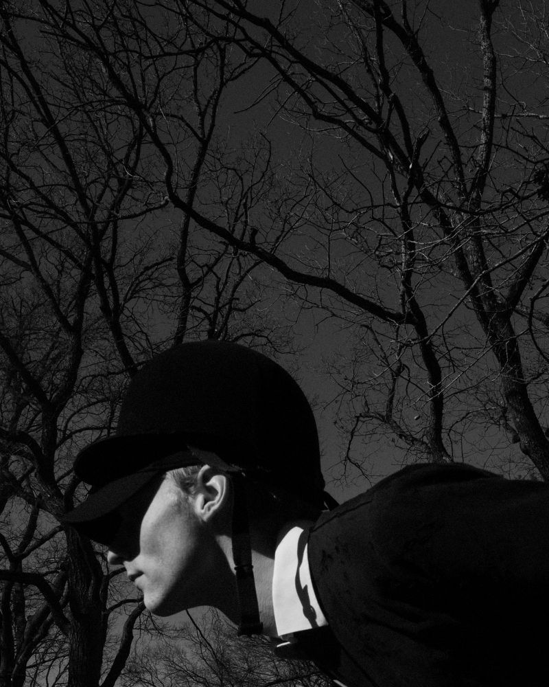 Black and white portrait of a person wearing equestrian helmet and dark riding jacket, with bare trees in the background.