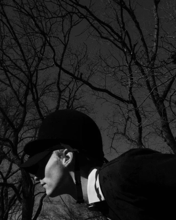 Black and white portrait of a person wearing equestrian helmet and dark riding jacket, with bare trees in the background.