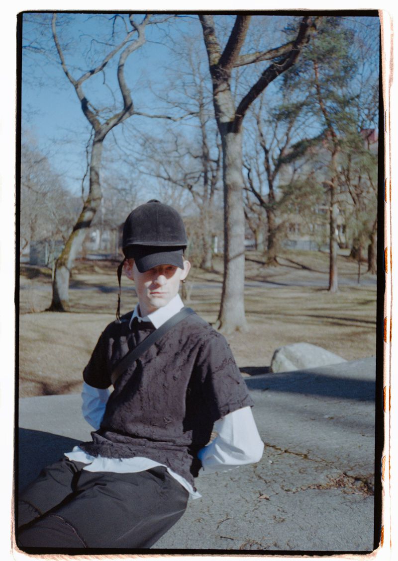 An adult male sits on a concrete surface outside wearing a black cap, dark shirt, white undershirt, dark pants, and a crossbody bag.