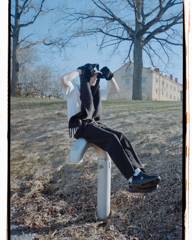 Young adult in a white t-shirt, black pants, and knit balaclava sitting on outdoor exercise equipment in a grassy, tree-filled park.
