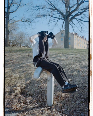 Young adult in a white t-shirt, black pants, and knit balaclava sitting on outdoor exercise equipment in a grassy, tree-filled park.