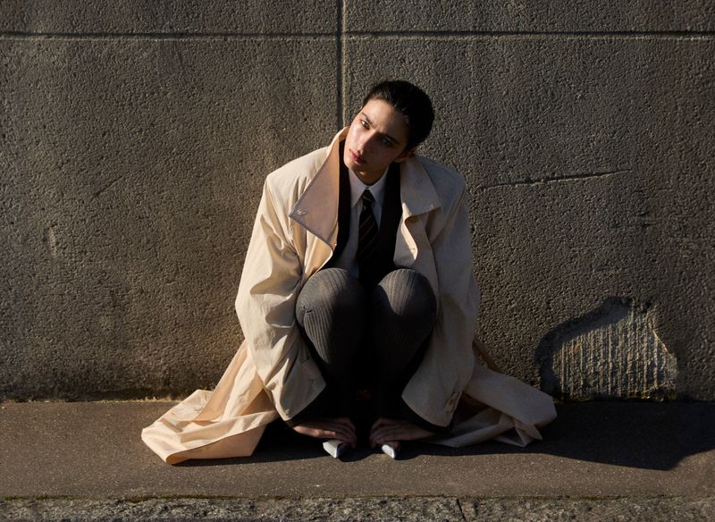Young adult in a trench coat squats on the ground outdoors, looking left with a concrete wall background in natural sunlight.