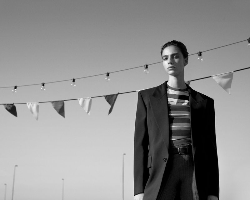 Black and white shot of a model in an oversized blazer and striped shirt, posing outdoors under pennant flags and clear sky.