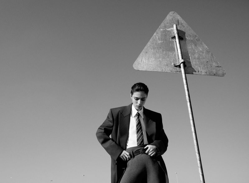 Monochrome portrait of a young adult wearing a suit, sitting near a triangle sign against a clear sky in an outdoor setting.