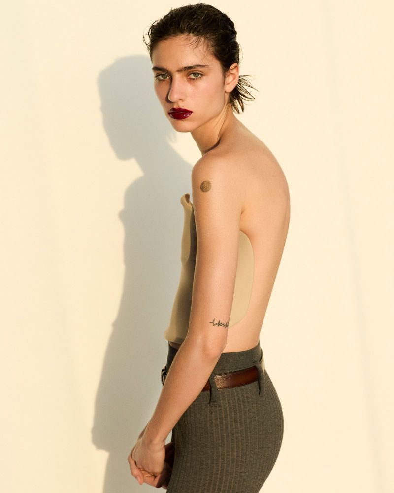 A young adult female poses indoors with a tan top and gray pants, showcasing a fashion editorial style, set against a cream background.