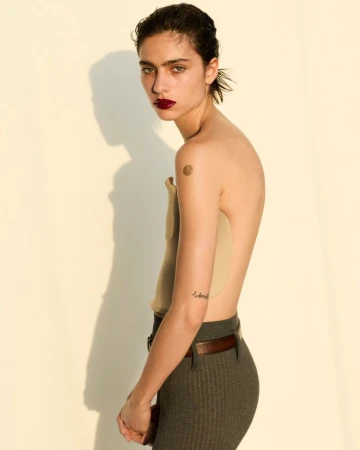 A young adult female poses indoors with a tan top and gray pants, showcasing a fashion editorial style, set against a cream background.