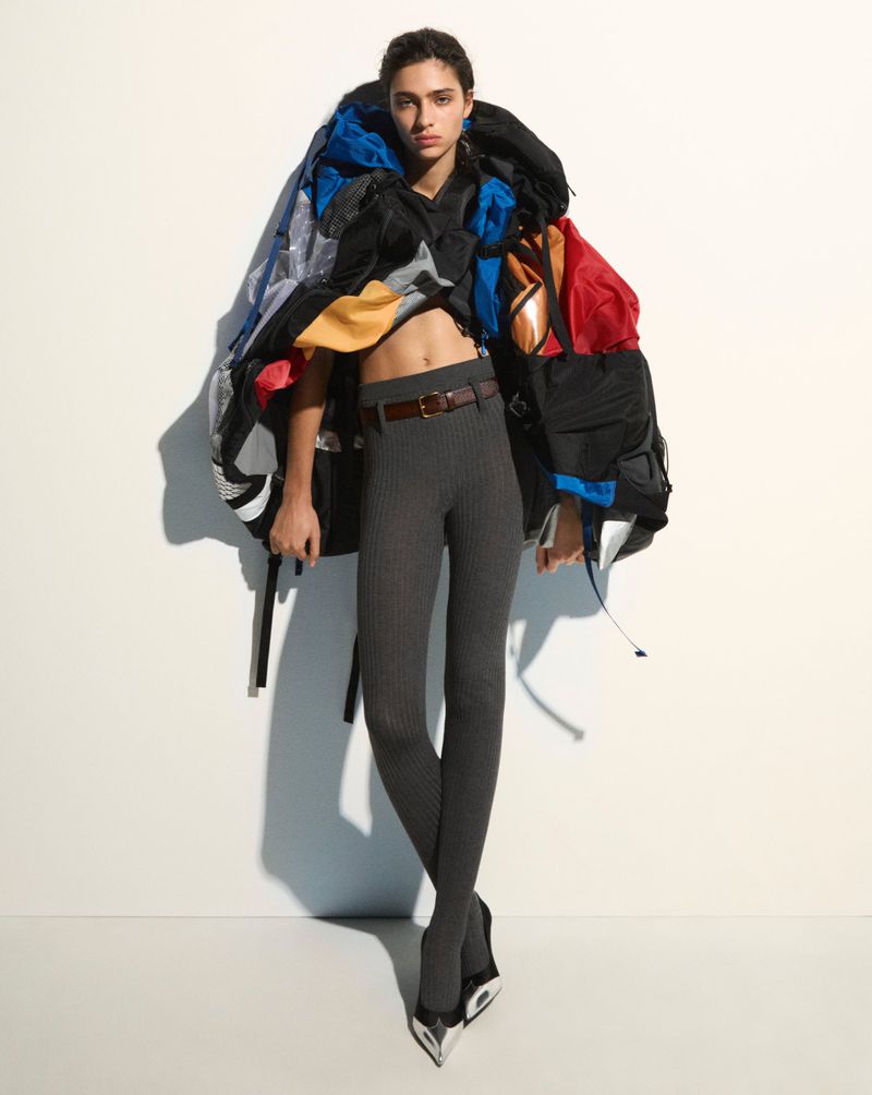Fashion-forward shot of a woman in grey leggings, black heels, and a colorful backpack-like garment against a stark white backdrop.