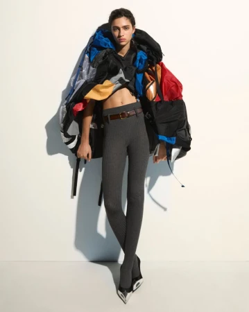 Fashion-forward shot of a woman in grey leggings, black heels, and a colorful backpack-like garment against a stark white backdrop.
