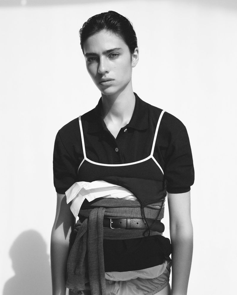Black and white portrait of a woman in a black shirt with layers of clothing and a belt wrapped around her waist, minimalist photo studio.
