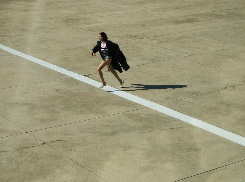 Woman in short shorts and heels runs on an empty tarmac wearing a black coat, her shadow cast on the concrete surface.
