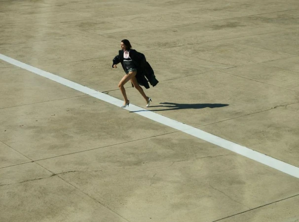 Woman in short shorts and heels runs on an empty tarmac wearing a black coat, her shadow cast on the concrete surface.