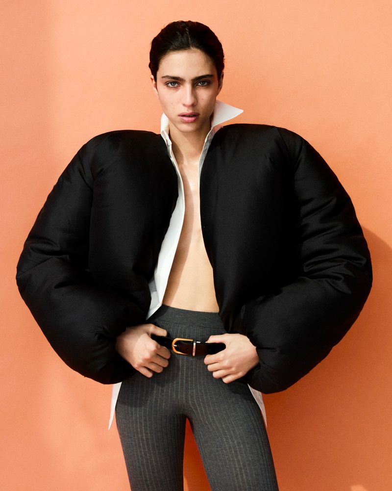 A fashionable adult female is posing in a photo studio wearing a black puffer jacket, grey leggings, and a white shirt, set against a peach background.