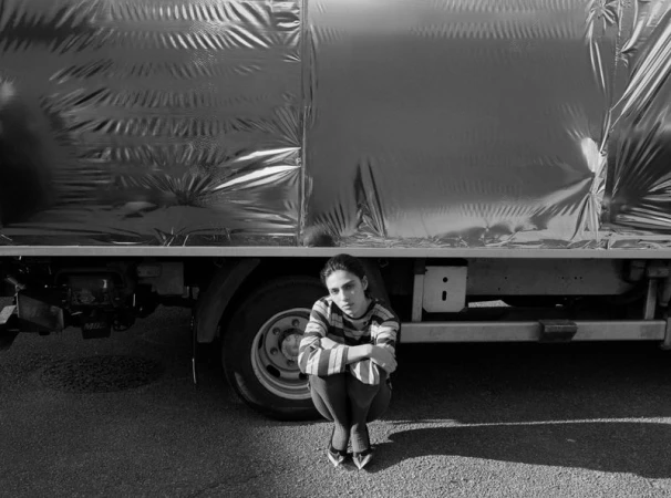 A young adult woman in striped shirt crouches in front of a truck covered in silver material, casting shadows on the asphalt ground.