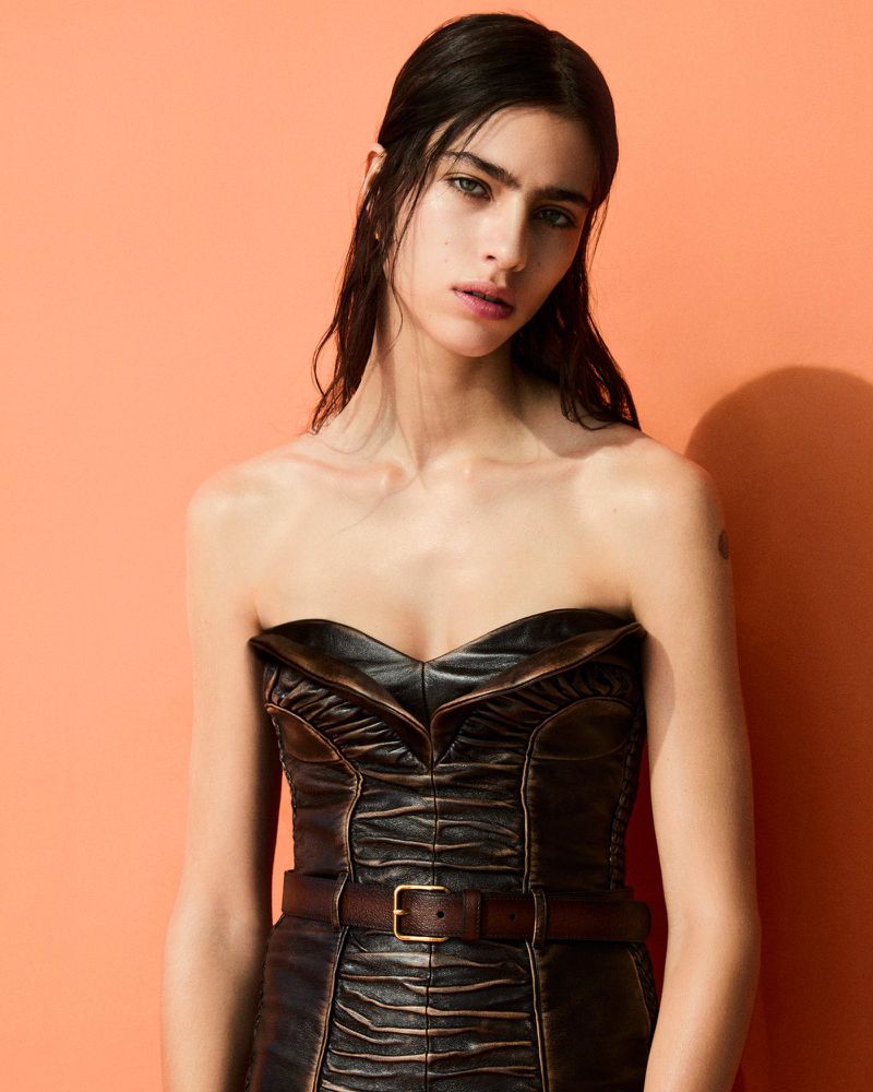 Brunette woman with fair skin poses indoors wearing strapless leather dress with brown belt against salmon-colored background in studio lighting.