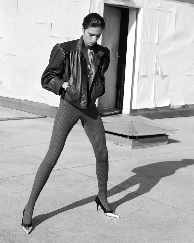 A stylish fashion shot in black and white features a model posing outdoors in a leather jacket, gray leggings, and heels against a building backdrop.