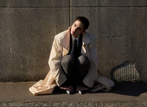 Woman in an oversized beige trench coat crouched against a textured stone wall