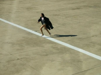Model running across large concrete pavement with a white line stripe, wearing a dark coat and high heels.