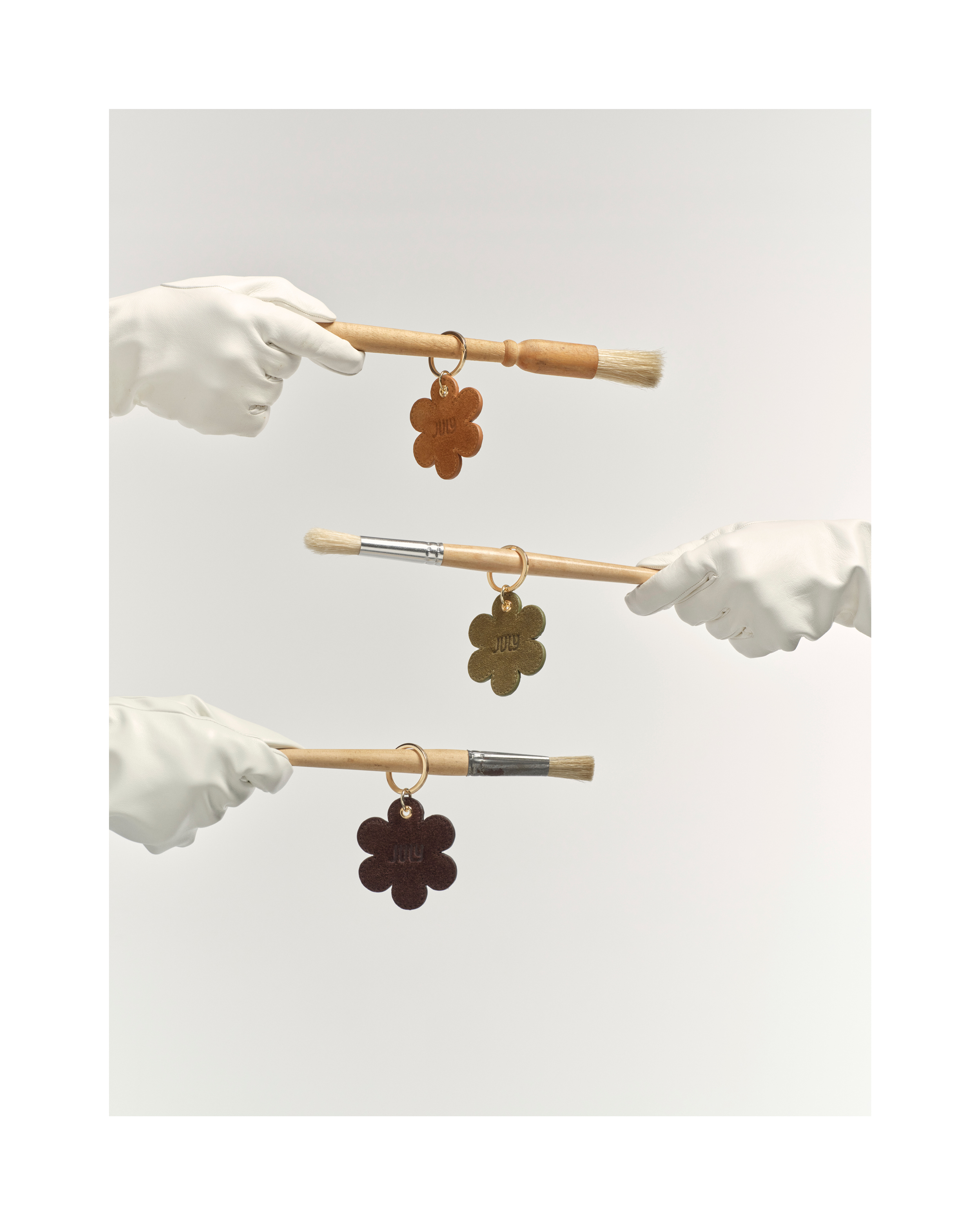 Three gloved hands holding paint brushes with flower-shaped keychains against a neutral background in a studio shot.