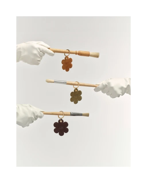 Three gloved hands holding paint brushes with flower-shaped keychains against a neutral background in a studio shot.