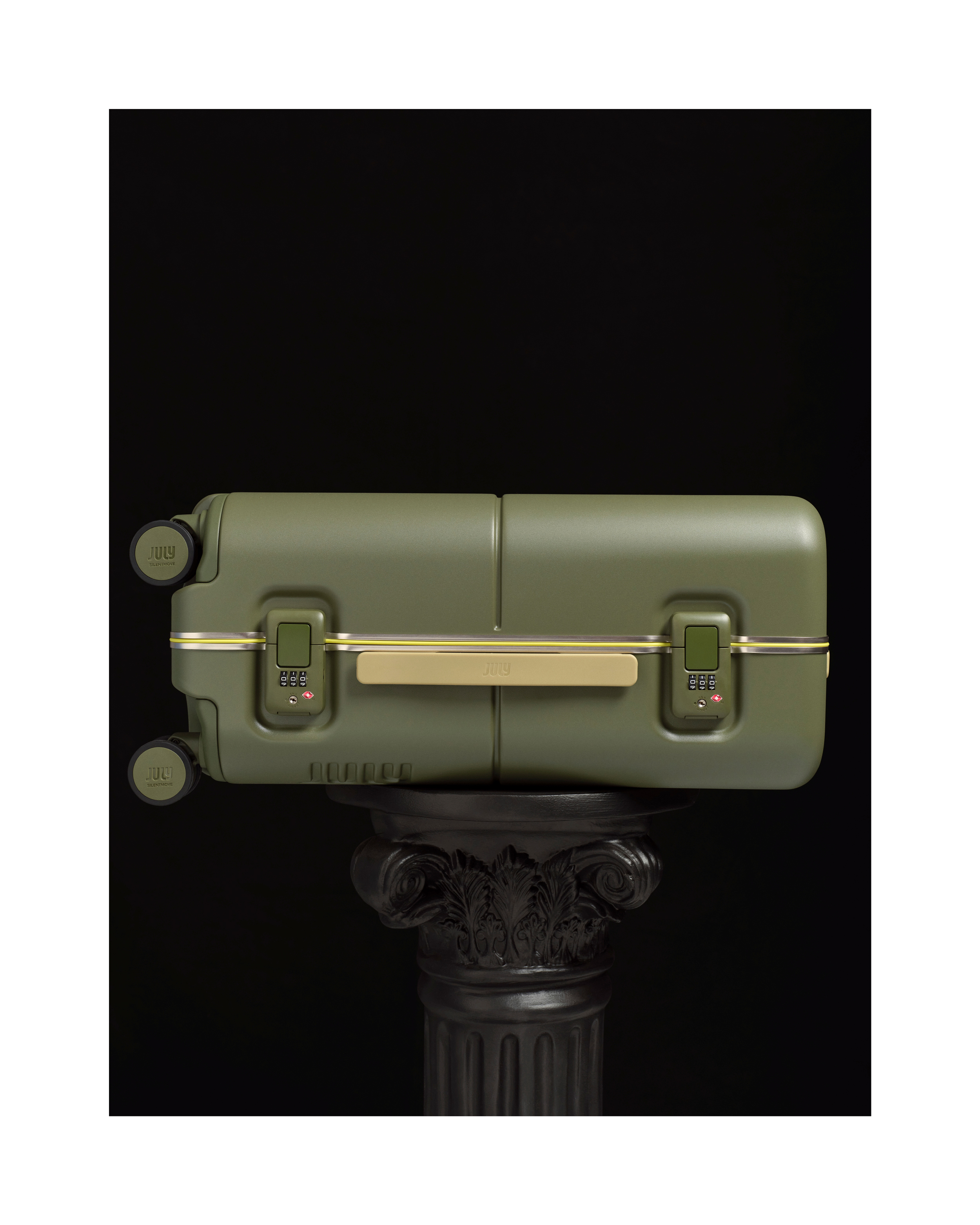 Olive green luggage closed and set sideways on black pillar in studio. Gold trim and locks detail the design of the suitcase.