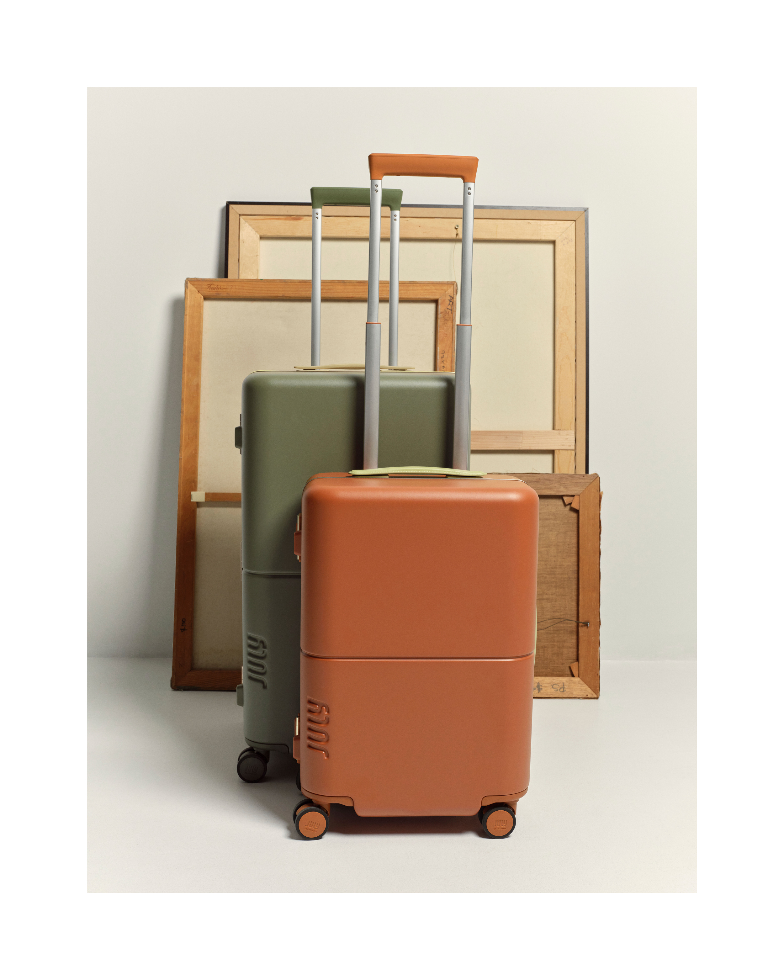 Studio shot of a pair of modern suitcases in burnt orange and sage green, arranged against a backdrop of framed canvases.