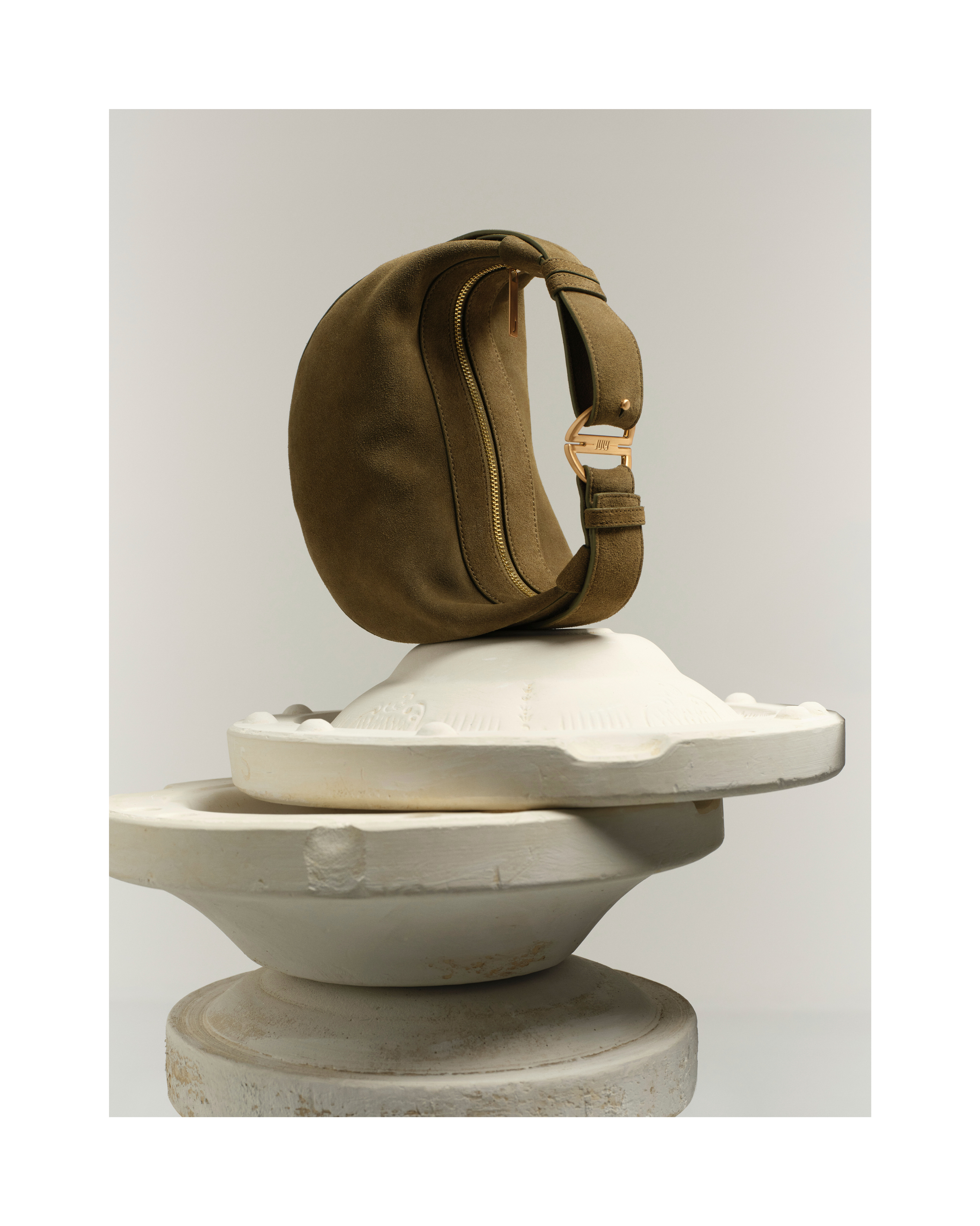 Olive green hobo bag with gold hardware displayed atop stacked white plaster forms, creating a minimalist fashion product still life.
