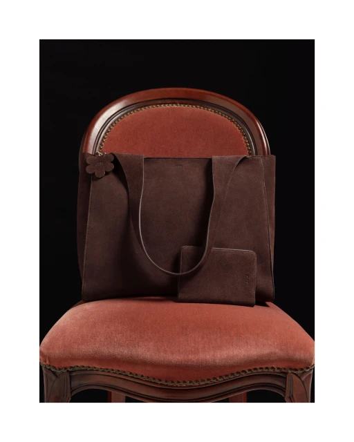 Brown suede tote bag and wallet set sitting on a vintage peach chair against a dark background, studio product shot, luxury.