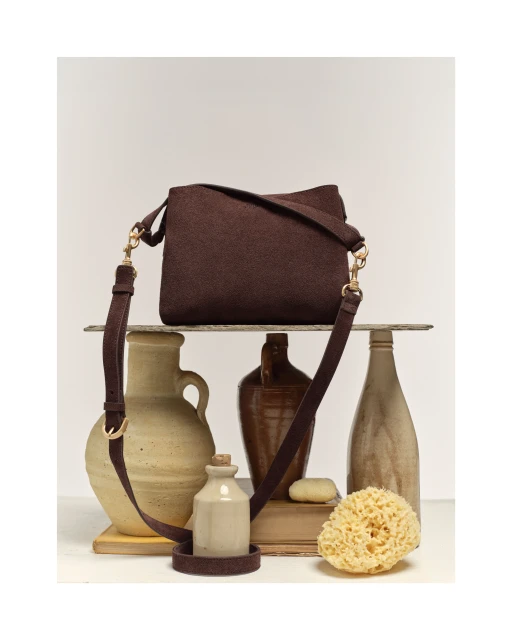 Studio shot of a brown textured crossbody bag with a brown strap, displayed with a collection of neutral colored ceramic vases.