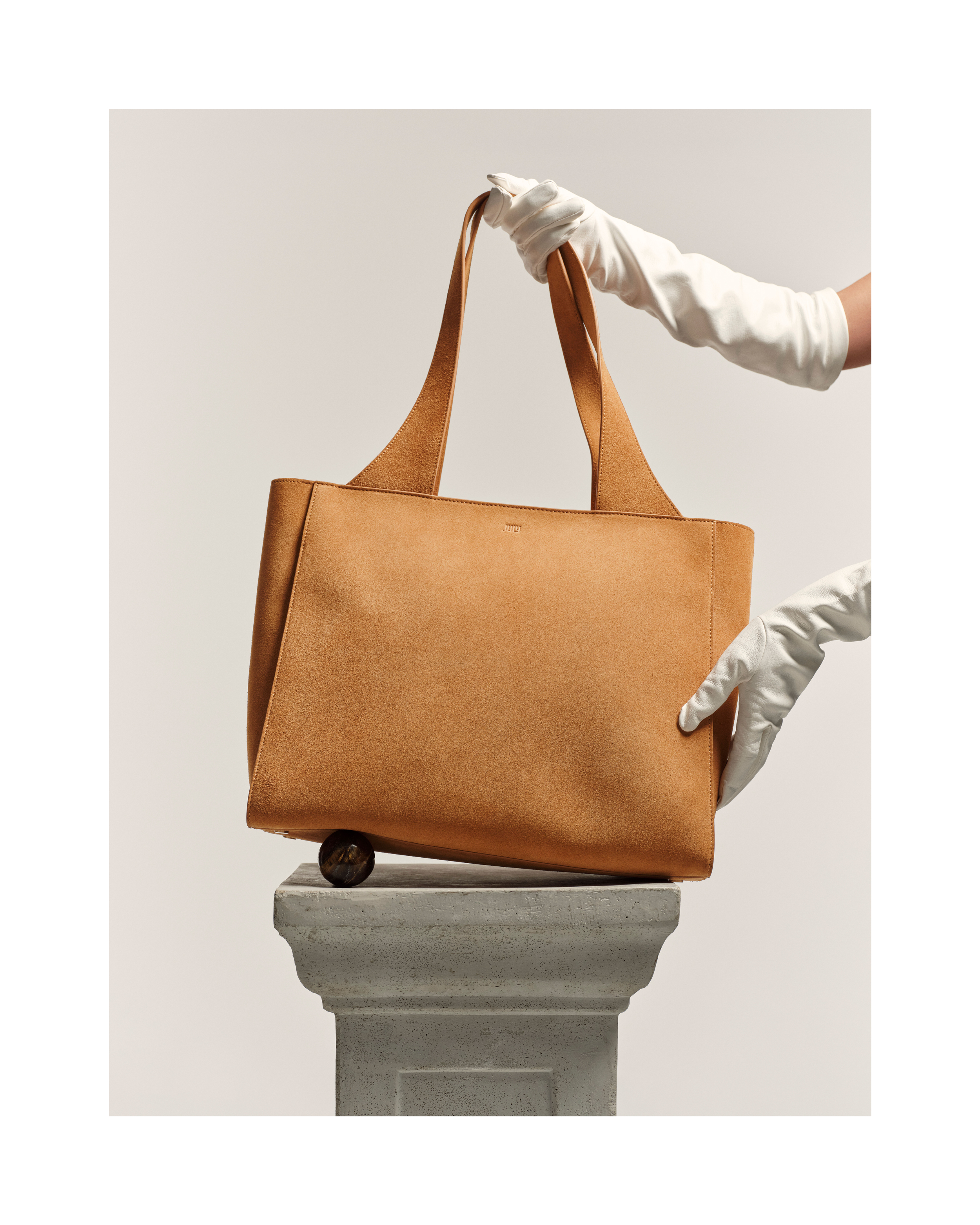 Tan leather tote bag on a pedestal, held by two hands in white gloves against an off-white background, promoting fashion and style.
