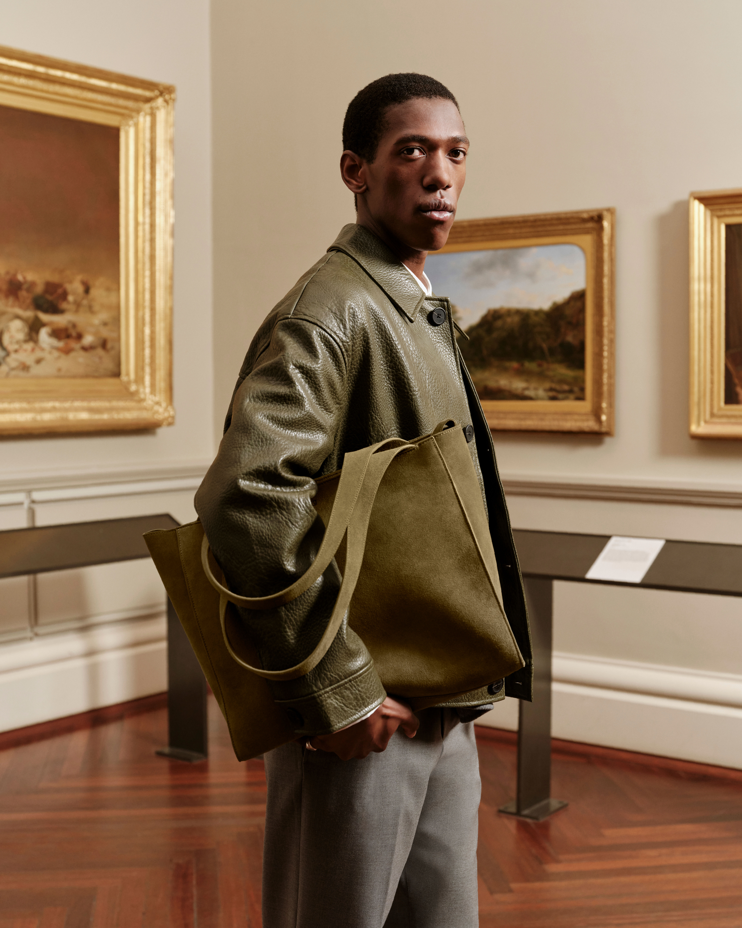 An adult male poses in a museum, wearing a green jacket and grey pants, carrying a green tote bag, with paintings in the background.