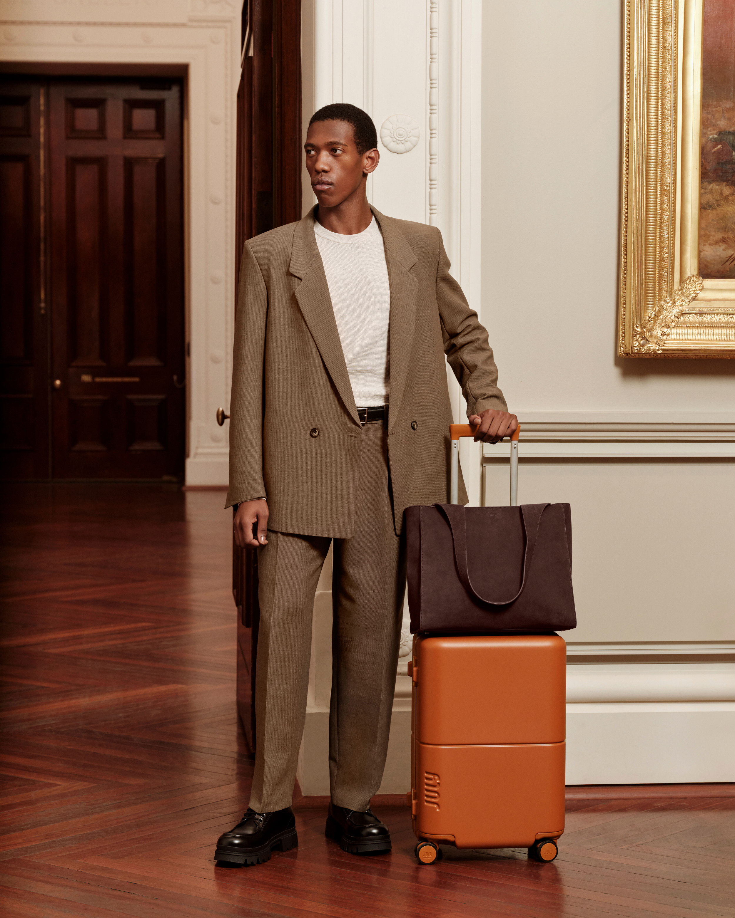 An African-American man in a brown suit with luggage in a building with hardwood floors, cream-colored walls, and a large painting.