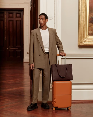 An African-American man in a brown suit with luggage in a building with hardwood floors, cream-colored walls, and a large painting.