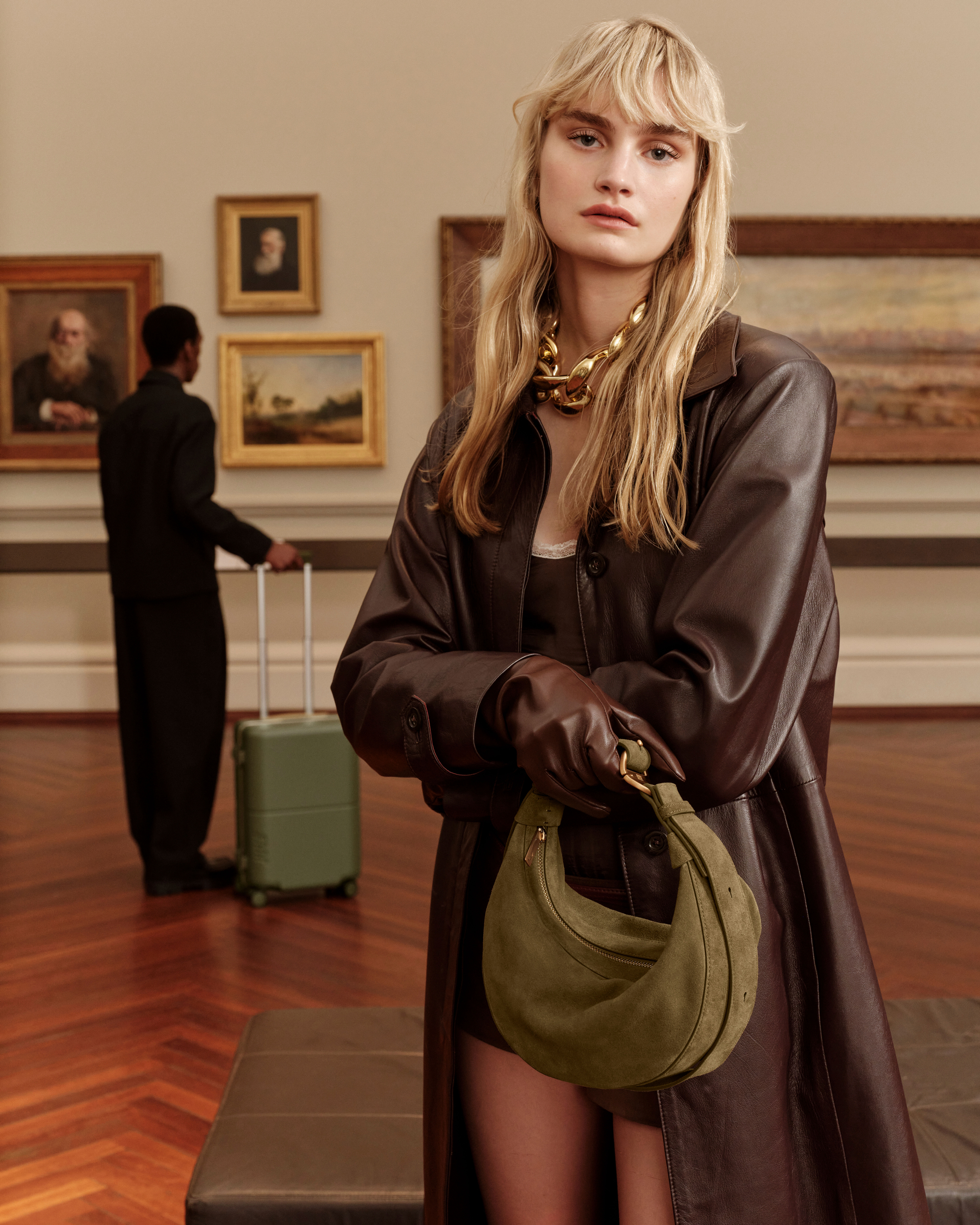 A blond female fashion model poses inside an art gallery wearing a brown coat, gold necklace, and green purse, with artwork in the background.