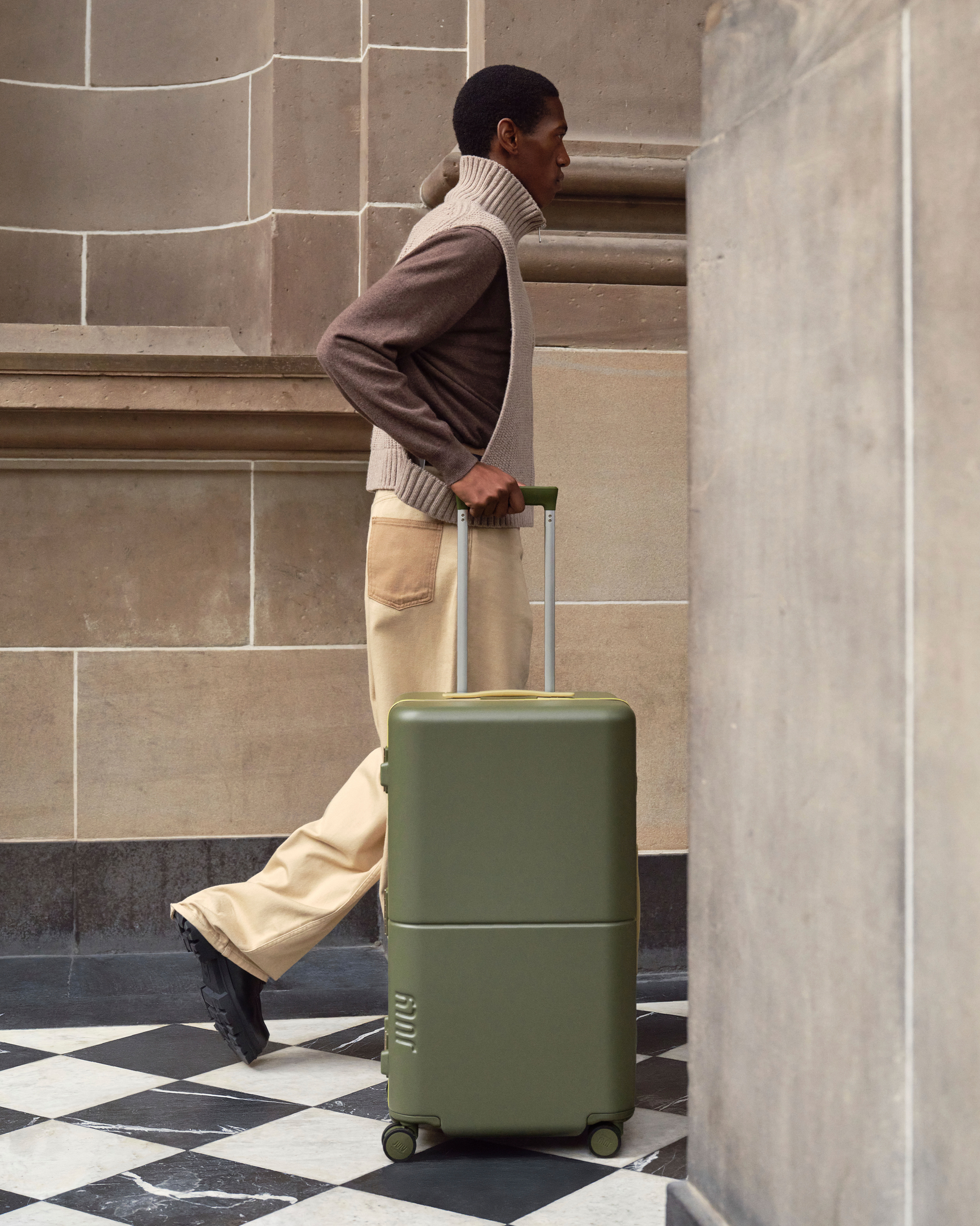 Stylish adult male walking with an olive-green suitcase in a neutral-toned architectural space with a black-and-white checkered floor.