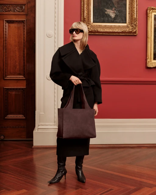 Blonde woman in a black coat and sunglasses holds a brown leather tote bag indoors in front of a red wall and dark wooden door.