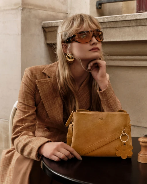 Adult woman wearing sunglasses and a brown blazer next to a yellow handbag with a flower keychain at an outdoor cafe table.