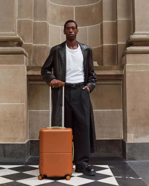 A Black man with a suitcase stands indoors against a stone wall, wearing a white t-shirt and a long black leather coat.