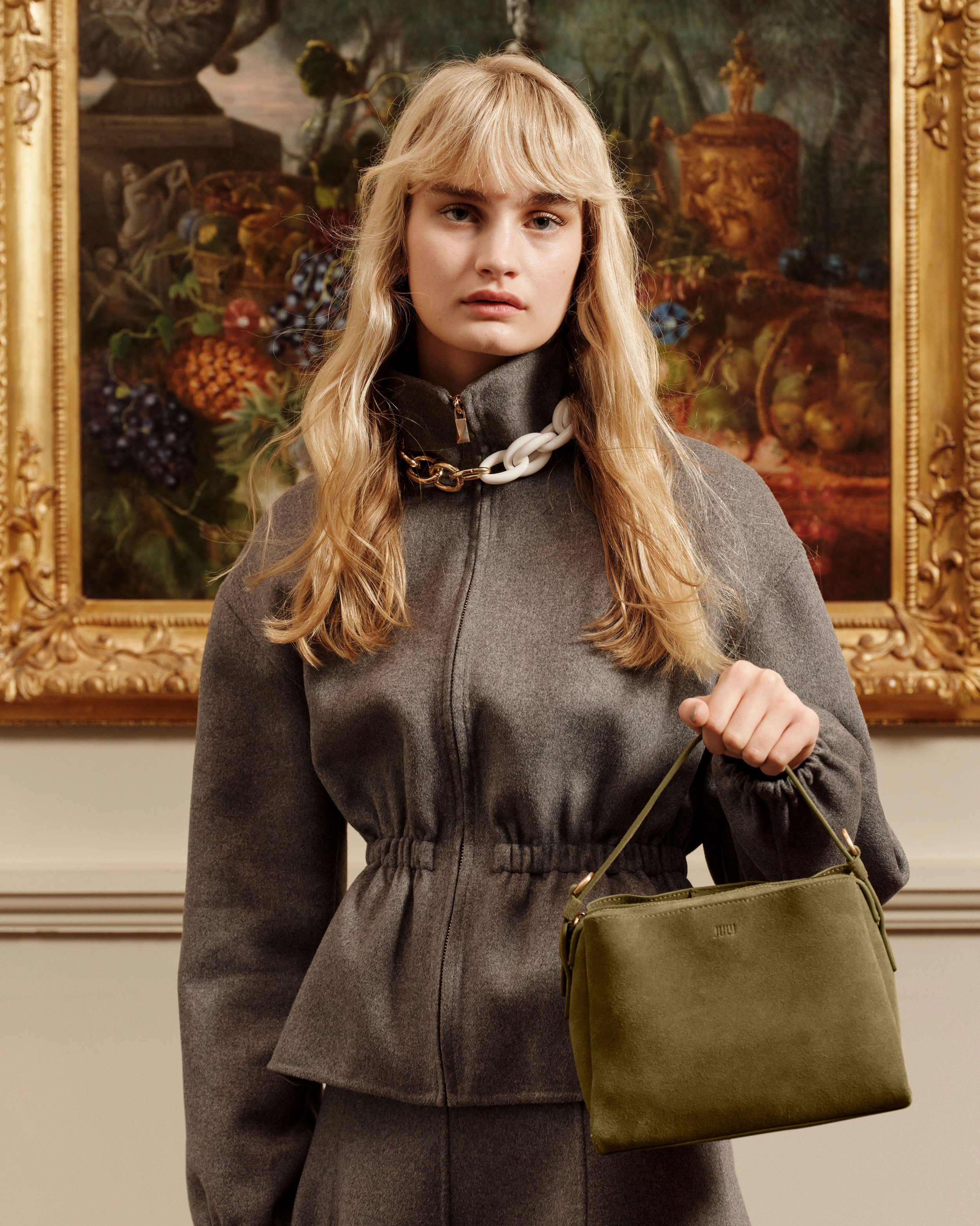 Woman wearing gray jacket and skirt holding green purse in an indoor setting with artwork; fashion and style product shot.