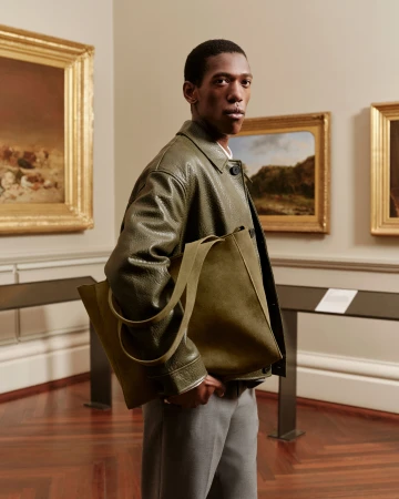Man wearing a green leather jacket holding an olive tote bag inside a museum gallery with framed paintings.