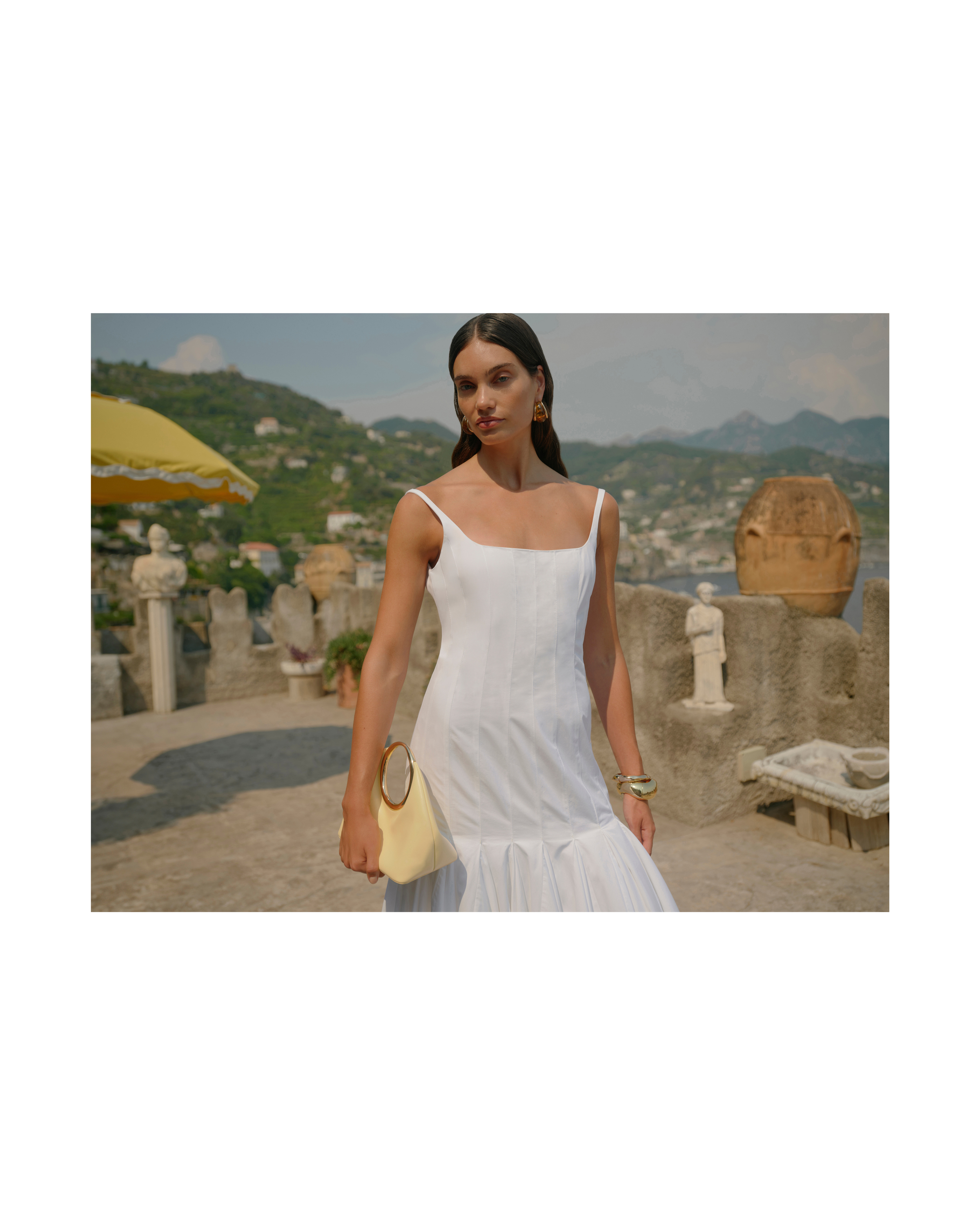 Woman in white dress poses outdoors on terrace with architecture, trees, and mountains visible; she holds a light yellow purse.