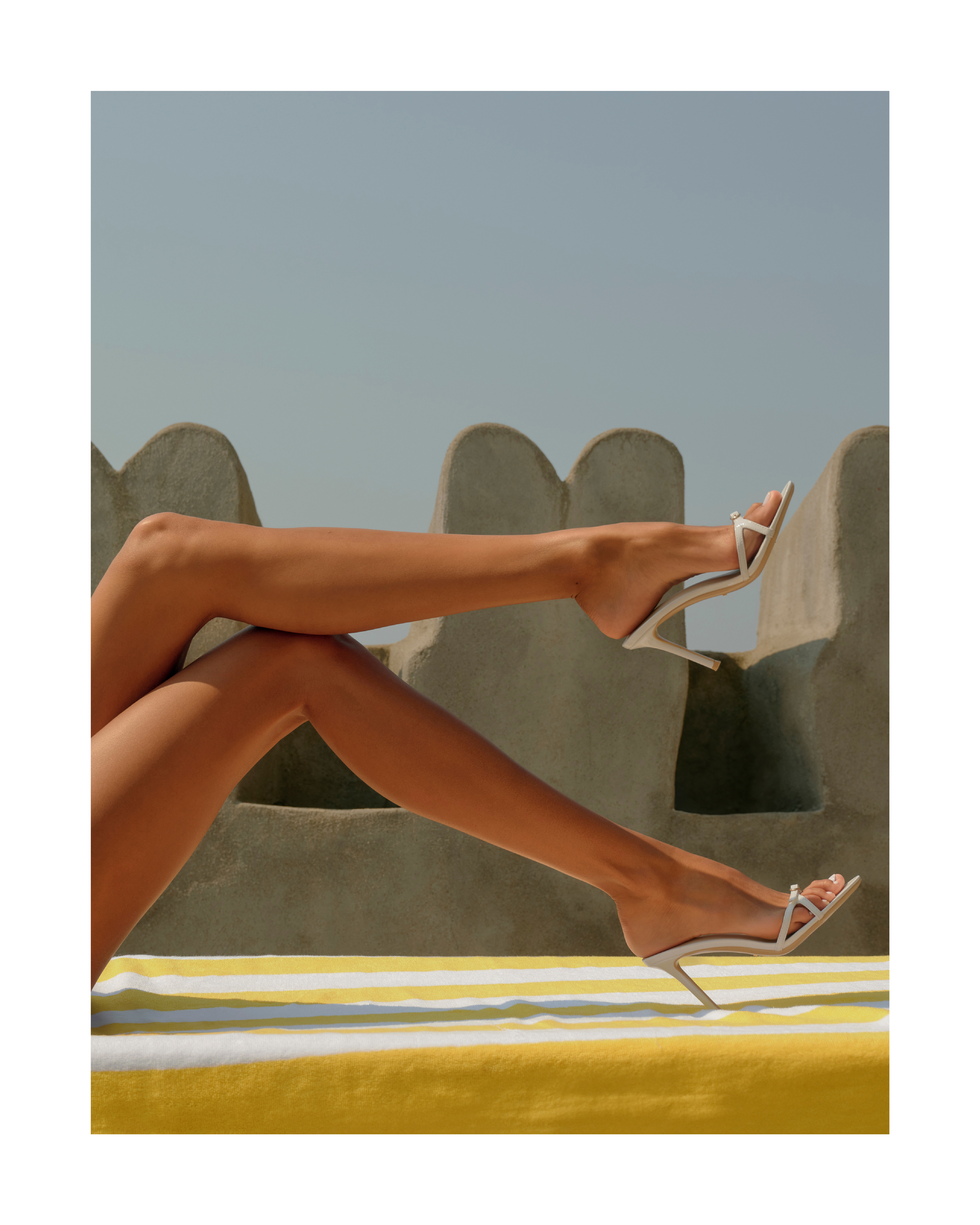 Woman's legs wearing strappy white high heels on a yellow and white striped surface with a gray wall and blue sky in the background.