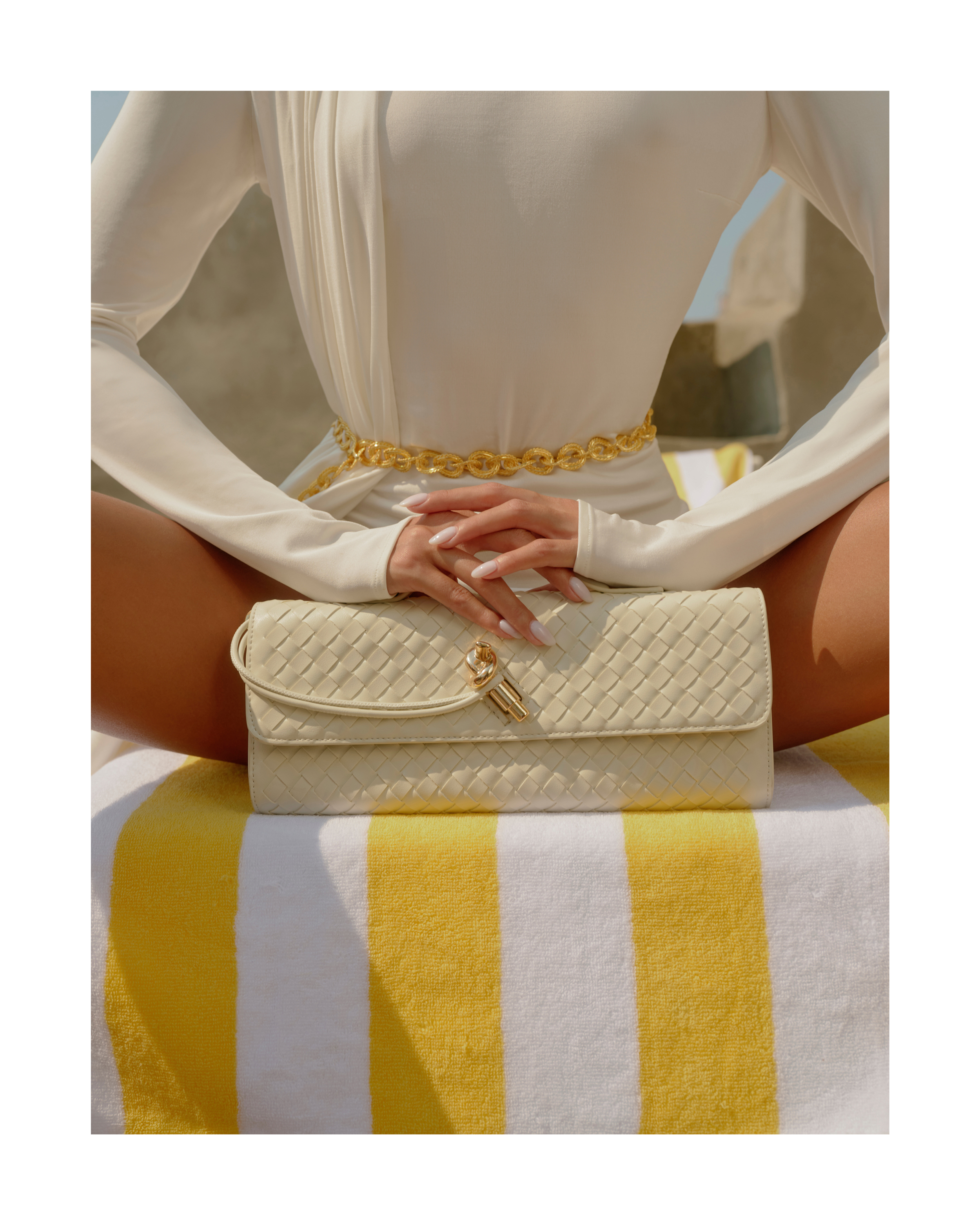 Woman in a white top and gold chain belt seated cross-legged holding a cream woven clutch bag on a yellow striped towel.