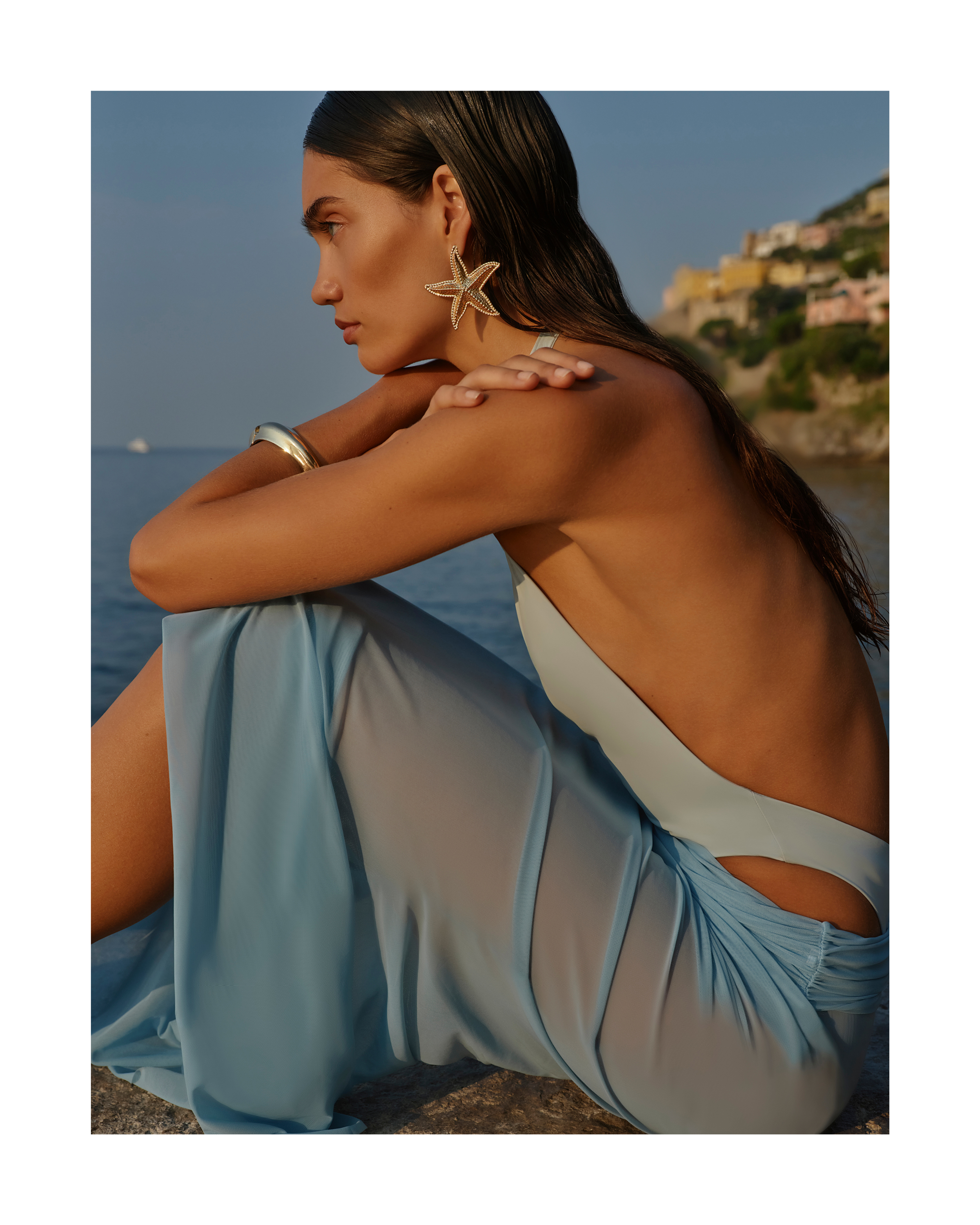 Woman in light blue dress sits with back exposed wearing starfish earrings and silver bracelet, with rocky coast and blue sea background.