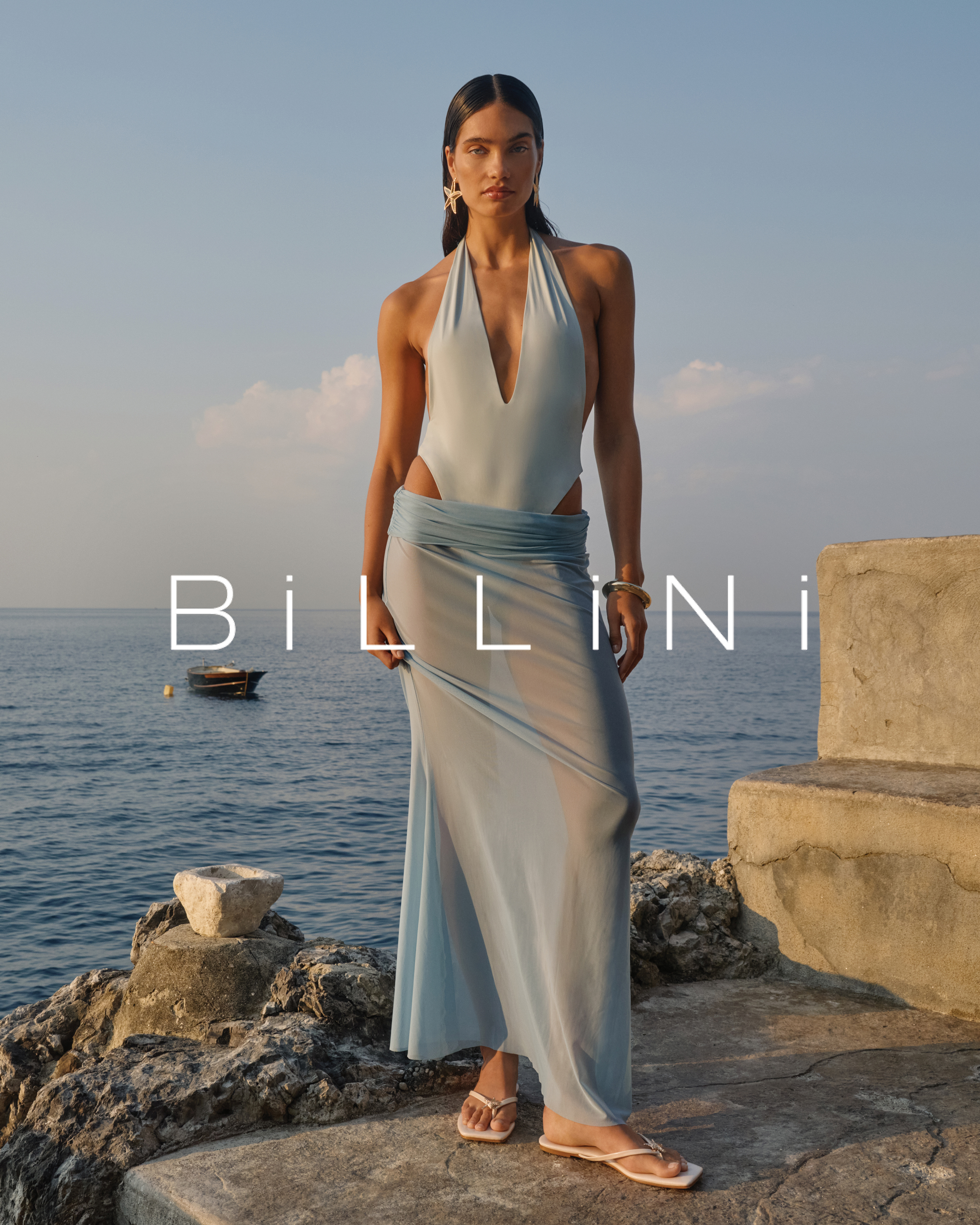 Adult female fashion model in a blue swimsuit with a sheer skirt stands on a stone ledge near the ocean in an outdoor setting.