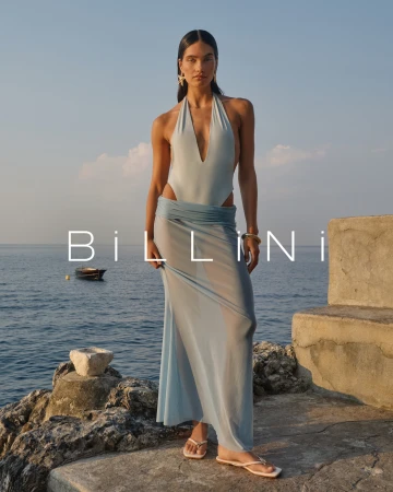 Adult female fashion model in a blue swimsuit with a sheer skirt stands on a stone ledge near the ocean in an outdoor setting.