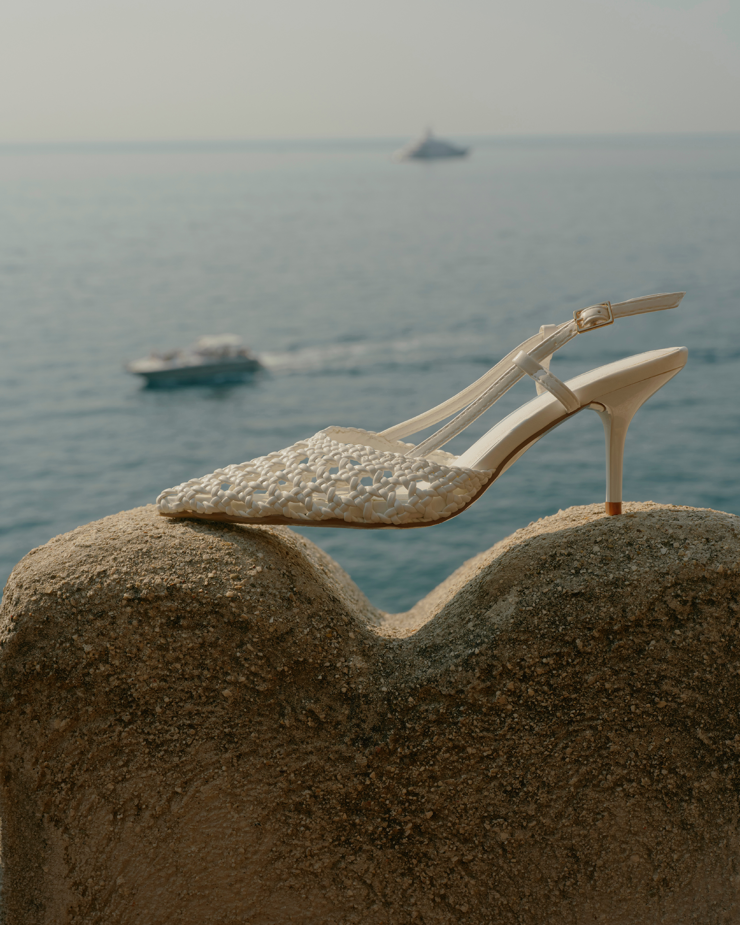 Elegant women's white woven slingback heel sits atop a sandy textured surface, with a blurred ocean and boat background.