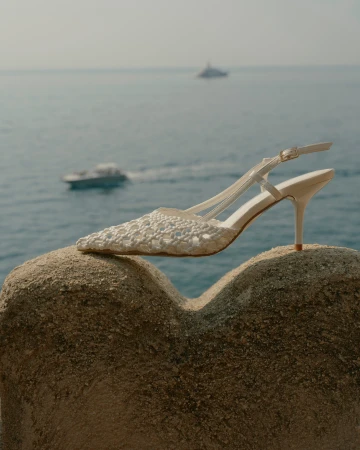 Elegant women's white woven slingback heel sits atop a sandy textured surface, with a blurred ocean and boat background.