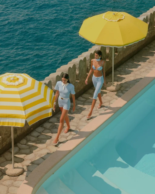 Two women in light blue outfits walk along a pool next to the ocean under bright yellow sun umbrellas, creating a summery vibe.